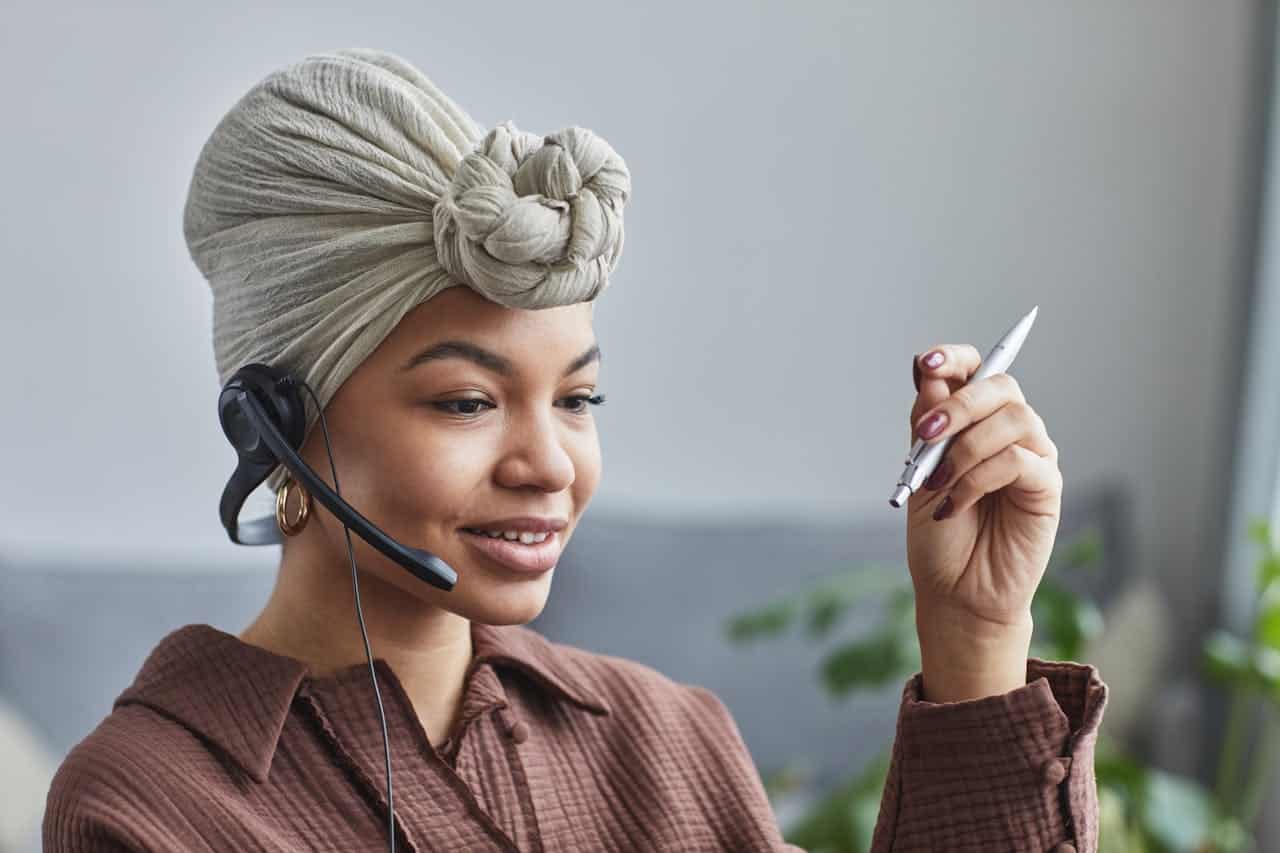 Operatrice call center con cuffia e microfono mentre lavora da remoto, penna in mano e sorriso professionale.